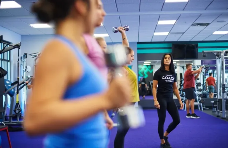 People exercising in a gym with equipment and a person wearing a "COACH" t-shirt in focus.